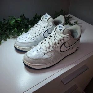 Nike Air Force 1 vita sneakers med blå detaljer - Klassiska vita Nike Air Force 1 sneakers med blå swoosh och blå yttersula. Skorna har snörning, rund tå och är tillverkade i skinn. Perfekta för dig som gillar en tidlös och sportig stil.