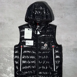 Svart dunväst med huva från Moncler - Snygg svart dunväst från Moncler med glansig finish och avtagbar huva. Västen har två framfickor med dragkedja och en bröstficka med Moncler-logga. Perfekt för lager-på-lager och håller dig varm under kyliga dagar.