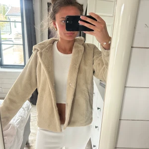 Beige teddyjacka med huva - Mysig beige teddyjacka med huva och knappar framtill. Jackan har en avslappnad passform och är perfekt för kyligare dagar. Den är fluffig och mjuk, vilket gör den extra skön att bära.