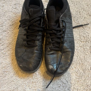 Svarta fotbollsskor från Puma - Säljer ett par svarta fotbollsskor från Puma i storlek 38. Skorna har snörning, rund tå och platt sula. De är tillverkade i syntetmaterial och har en diskret mönstrad yta. Perfekta för dig som vill ha stilrena skor på planen.