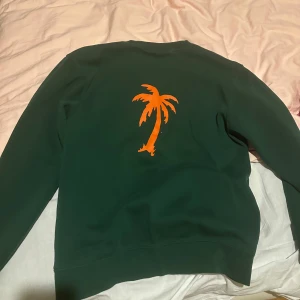 Grön sweatshirt från Mucker med palm - Mörkgrön sweatshirt från Mucker med orange tryck framtill och en stor orange palm på ryggen. Tröjan har rund hals, långa ärmar och är gjord i mjuk bomullsmix. Perfekt för dig som gillar streetwear och vill sticka ut med coola detaljer.