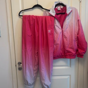 Adidas set, jacka med tillhörande byxor (Primegreen) - Supercoolt tvådelat set från Adidas Originals i ombre färgskiftning från rosa till vit. Både jackan och byxorna är i lätt, prasslig sportmaterial. Perfekt för streetwear, festival eller vardag med attityd. Finns inte längre att köpa på adidas hemsida. 