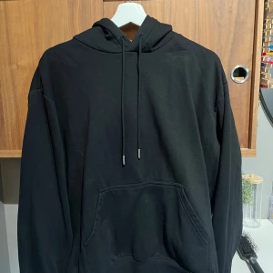 Svart hoodie med huva och magficka - En klassisk svart hoodie med huva och snörning samt en stor magficka framtill. Tröjan har lång ärm och en relaxed passform, perfekt för chill dagar. Materialet känns mjukt och skönt, troligtvis bomull eller en bomullsblandning.