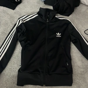 Svart Adidas flis med vita ränder - Säljer en svart Adidas flis med klassiska vita ränder längs ärmarna och stor vit trefoil-logga på ryggen. Flisen har dragkedja framtill, hög krage och två fickor med dragkedja. Perfekt för en sportig och stilren look.