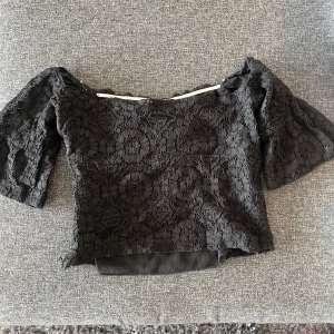 Svart offshoulder spetsblus - Säljer en svart blus med offshoulder-modell och trekvartsärmar. Toppen är täckt av vacker spets med blommigt mönster och har en croppad passform. Perfekt till fest(Ai😁)