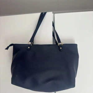 Mörkblå  axelväska med dragkedja - Stilren mörblå axelväska med långa remmar och guldfärgade detaljer. Väskan är rymlig med flera innerfack och stängs med dragkedja. Perfekt för dig som vill ha plats för allt du behöver under dagen. Den har inga defekter. Men eftersom den legat i en låda ett bra tag är den lite skrynklig. Väskan är helt ny och har aldrig använts. Pris kan diskuteras