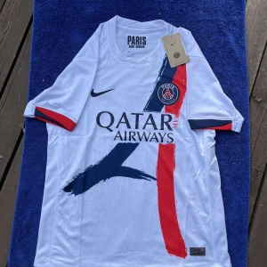 PSG vit bortatröja Nike 2023/24 - Paris Saint-Germain bortatröja i vitt med blå och röd penseldrag-design på framsidan. PSG-emblem på bröstet, Nike-logga och Qatar Airways sponsortryck. Tillverkad i lätt Dri-FIT-material som andas. Kortärmad modell med blå och röda detaljer på ärmsluten.