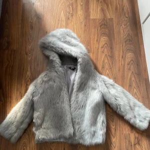 Grå pälsjacka med huva, strl 32 - Supermjuk och fluffig grå pälsjacka med huva. Jackan har lång ärm och är helfodrad med silkeslen insida. Perfekt för dig som vill sticka ut och hålla dig varm med stil. Snygg oversized känsla och passar till många olika looks.