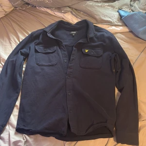 Mörkblå skjorta från Lyle & Scott Junior - Mörkblå skjorta från Lyle & Scott Junior med klassisk krage, två bröstfickor med lock och knappar samt den ikoniska gula loggan på vänster ficka. Skjortan är långärmad och tillverkad i 100% bomull för en skön känsla. Passar folk som är runt 170 Bra skick kom med frågor