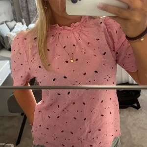 Rosa blommig blus med volangkrage - Supersöt rosa blus med broderade blommor och hålmönster. Volangdetalj vid halsen och korta puffärmar ger en gullig vibe. Perfekt för dig som gillar romantisk stil och vill sticka ut lite extra.