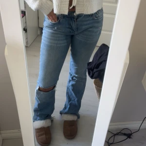 Blå bootcut jeans med hål på knät - Säljer ett par ljusblå jeans med bootcut-modell och snyggt hål på ena knät. Jeansen har klassisk femficksdesign, normal passform och är tillverkade i jeansmaterial. Perfekta för dig som gillar en avslappnad och trendig look.