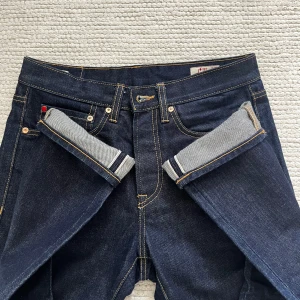 SELVAGE jeans från 157 Roots - Sjukt snygga mörkblå selvage jeans från 157 Roots, modell R003. Klassisk rak passform med orange kontrastsömmar, knäppning med knappar och detaljerad selvage-kant. Rå denim i 14,5 oz bomull med 3x1 right hand twill. Perfekta för dig som gillar stilrena jeans med edge. Det är bara att höra av sig vid frågor 🙌