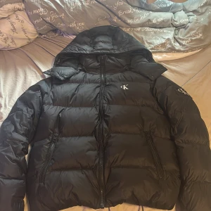Svart pufferjacka från Calvin Klein Jeans - Säljer en svart pufferjacka från Calvin Klein Jeans med huva och dragkedja framtill. Jackan har två snedställda fickor med dragkedja och diskret CK-logga på bröstet och ärmen. Yttermaterialet är återvunnen nylon och fodret är återvunnen polyester. Nypris 2699kr använd fåtal gånger mycket bra skick pris går att diskuterat och kom med frågor