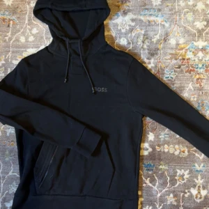Svart hoodie från BOSS med huva - Snygg svart hoodie från BOSS med diskret logga på bröstet. Tröjan har huva med snörning och en stor magficka framtill. Perfekt för en chill och stilren look. Långärmad och i mjukt material som känns skönt mot huden. Nypris 1300 Zalando