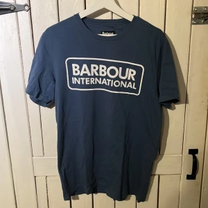 Blå Barbour International t-shirt - Snygg blå t-shirt från Barbour International med stort vitt logotryck på bröstet. Klassisk rund hals och korta ärmar. Perfekt för dig som gillar stilrena och bekväma plagg med tydlig branding.