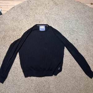 Mörkblå stickad tröja Lindbergh - Snygg mörkblå stickad tröja från Lindbergh med relaxed fit. Tröjan har ett klassiskt kabelstickat mönster och långa ärmar. Perfekt för dig som gillar enkel och stilren design med lite extra struktur. Passar till jeans eller chinos för en chill look. Väldigt relaxad stil