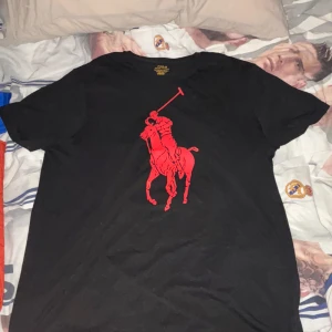 Svart t-shirt Polo Ralph Lauren - Svart t-shirt från Polo Ralph Lauren med stor röd polospelare tryckt på bröstet. Klassisk rund hals och korta ärmar. Snygg och enkel design som sticker ut med den ikoniska loggan. Modellen är custom slim fit och materialet är mjuk bomull. Priset kan alltid diskuteras 