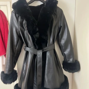 Svart pälsfodrad skinnjacka med bälte - Snygg svart skinnjacka med mjukt pälsfoder och pälsdetaljer på huva, ärmslut och nederkant. Jackan har bälte i midjan för en schysst siluett och är perfekt för kalla dagar. Långärmad och med en cool, lite oversized vibe. Den är använd bara en gång men den är helt ny inga skador eller någonting. 