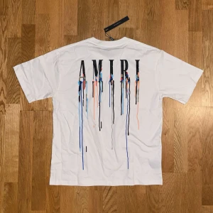 Vit AMIRI t-shirt med färgstänk - Vit t-shirt från AMIRI med coolt tryck av loggan och färgstänk i blått, svart och orange på både bröstet och ryggen. Klassisk passform med rund hals och korta ärmar. Perfekt statement-plagg för dig som gillar streetstyle.