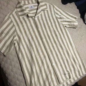 Randig kortärmad skjorta från NN07 - Snygg kortärmad skjorta från NN07 i beige och vit med breda vertikala ränder. Skjortan har klassisk krage, bröstficka och knäppning framtill. Tillverkad i ett strukturerat linnematerial som känns luftigt och perfekt för varma dagar. Fits medium sällan använd max 4 ggr
