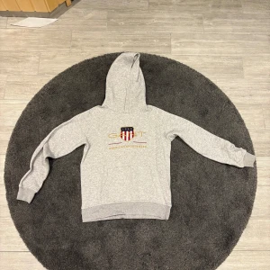 Grå hoodie från GANT med broderad logga - Snygg grå hoodie från GANT med broderad logga och sköld på bröstet. Tröjan har huva, långa ärmar och ribbade muddar. Perfekt för en chill och sportig look. Klassisk design med amerikansk sportswear-vibe.