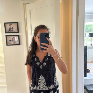 Mörkblå broderad linnetopp Indiska - Supersöt mörkblå linnetopp från Indiska med vita broderade blommor och snygg v-ringning. Toppen är ärmlös och har en avslappnad passform, perfekt för varma dagar. Broderidetaljerna ger en bohemisk vibe och toppen är gjord i mjuk bomull.