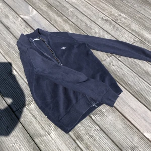 Mörkblå stickad kofta med dragkedja - Mörkblå stickad kofta med hel dragkedja framtill och ribbade muddar. Klassisk och enkel design med liten broderad logga på bröstet. Perfekt för lager-på-lager under kyliga dagar.