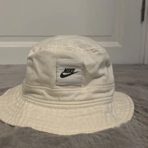 Vit bucket hat från Nike - Snygg vit bucket hat från Nike med klassisk logga framtill. Hatten är tillverkad i bomull och har en avslappnad passform med bred brätte. Perfekt accessoar för att lyfta din streetwear-look eller skydda dig mot solen.