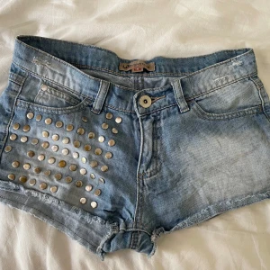 Ljusblå jeansshorts med nitar - Snygga ljusblå jeansshorts från LAB Industries med coola silvriga nitar på ena framsidan. Klassisk femficksmodell, råa kanter och slitningar för en edgy look. Perfekta för sommardagar när du vill sticka ut lite extra.