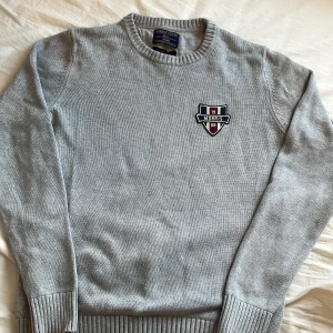 Grå stickad tröja från Morris Gents - Snygg grå stickad tröja från Morris Gents i storlek medium. Tröjan har rund halsringning, ribbade muddar och ett broderat Morris-emblem på bröstet i marinblått, vinrött och vitt. Perfekt för dig som gillar klassisk och stilren look. Storlek m men sitter som s