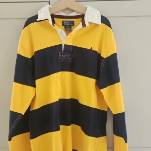 Randig pikétröja Polo Ralph Lauren - Gul och marinblå randig pikétröja från Polo Ralph Lauren med klassisk vit krage och lång ärm. Tröjan är i 100% bomull och har broderad logga på bröstet. Perfekt för dig som gillar sportig och preppy stil.