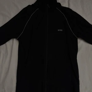Svart zipjacka från BOSS, stl S - Svart zipjacka från BOSS med broderad logga på bröstet och vita kontrastsömmar längs axlarna. Jackan har huva, hel dragkedja och två fickor framtill. Perfekt för en clean och sportig look.