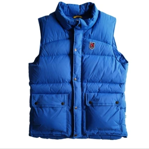 Blå dunväst från Fjällräven - Säljer en sådan här blå dunväst från Fjällräven med hög krage och två stora fickor framtill. Västen har tryckknappar och klassisk Fjällräven-logga på bröstet. Perfekt att slänga över en hoodie för extra värme.