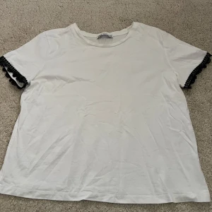Vit t-shirt med spetsdetalj från Zara - Vit t-shirt från Zara i bomull med korta ärmar. Ärmsluten har svarta spetsdetaljer som ger en snygg kontrast. Klassisk rund halsringning och rak passform. Perfekt för dig som gillar enkla plagg med en twist.
