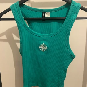 Turkos ribbad linnetopp från Divided - Säljer en turkos ribbad linnetopp från Divided i storlek S. Toppen är ärmlös och croppad med en broderad vit detalj framtill. Perfekt för dig som gillar en enkel men ändå unik look.