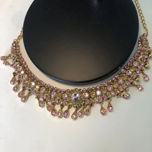 Guldigt halsband med rosa stenar - Statement-halsband i guldton med massor av glittrande rosa stenar och droppformade hängen. Perfekt för att sticka ut och ge din outfit extra bling. Kedjan är justerbar och smycket har en lyxig vintagekänsla.