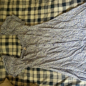 Blommig blå klänning från Nelly - Supersöt blå klänning från Nelly med vitt blommönster, kort ärm med volang och knappar framtill. Perfekt för sommarens alla tillfällen! Lätt och luftig modell som ger en romantisk vibe.