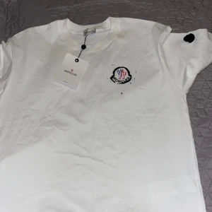 Vit Moncler t-shirt med tryck, M - Vit t-shirt från Moncler med coolt färgstarkt tryck i blått och rött på ryggen och mindre logga framtill. Klassisk rund hals, korta ärmar och svart moncler logga på ena ärmen. Skön bomullskvalitet och modern passform. Har kvitto 