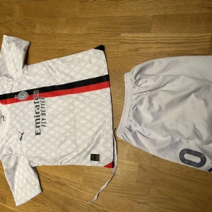 AC Milan bortatröja Leão #10 Puma - AC Milan bortaställ med Leão #10. Vit kortärmad tröja med subtilt geometriskt mönster, svarta och röda diagonala ränder, Puma-logga och sponsortryck. Matchande vita shorts med nummer. Perfekt för dig som älskar fotboll och vill sticka ut på planen.