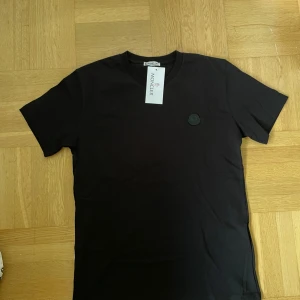 Moncler t shirt  - Svart t-shirt från Moncler med klassisk rund hals och diskret svart logomärke på bröstet. T-shirten är enkel och stilren, perfekt för dig som gillar minimalistisk design. Priset är inte hugget i sten