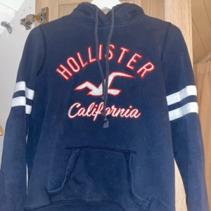 Marinblå Hollister hoodie med tryck - Snygg marinblå hoodie från Hollister med rött och vitt broderat tryck på bröstet och vita ränder på ärmarna. 