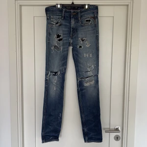 Blå slitna jeans från Replay - Säljer ett par blå jeans från Replay med rejält slitna detaljer och hål på både fram- och baksida. Jeansen har klassisk femficksdesign, raka ben och snyggt tvättad finish. Perfekta för dig som gillar en rå och avslappnad streetstil.