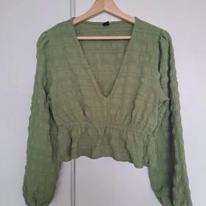 Olivgrön blus med v-ringning - Supersöt olivgrön blus med v-ringad hals och lång puffig ärm. Toppen har ett krinklat tyg med struktur, markerad midja och lätt volang nertill. Perfekt för dig som gillar en chill men ändå trendig look.