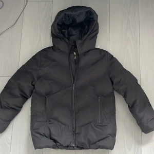Svart pufferjacka från Zara med huva - Säljer en svart pufferjacka från Zara i storlek 152. Jackan har en stor huva, två fickor med dragkedja och hel dragkedja framtill. Perfekt för kalla dagar och riktigt skön att bära. Enkel, stilren och passar till det mesta.