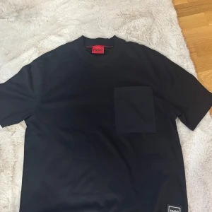 Hugo-tshirt - Svart t-shirt från Hugo med klassisk passform, rund halsringning och en stor bröstficka. T-shirten har en stilren look med diskret Hugo-logga nertill och är tillverkad i mjuk bomull. Perfekt för en clean och modern stil. Nypris:1200