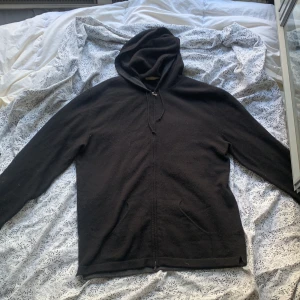 Svart hoodie från Luca Faloni i ull - Svart hoodie från Luca Faloni i mjuk ull. Klassisk modell med dragkedja, snörning i huvan och två fickor framtill. Perfekt för dig som gillar en clean och stilren look med premiumkänsla. Snygg att bära över en skjorta eller t-shirt.