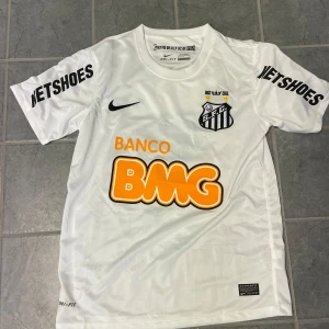 Ny Santos Neymar Jr #11 Nike fotbollströja - Vit Santos fotbollströja från Nike med Neymar Jr och nummer 11 på ryggen. Tröjan har korta ärmar, svarta tryck och orange BMG-logga. Tillverkad i Dri-Fit-material som andas, med klubbmärke och sponsorer på bröst och ärmar.