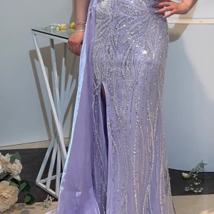 Ljuslila glittrig balklänning med slits - Magisk långklänning i ljuslila – använd endast i tre timmar!  En riktigt elegant och unik klänning med glittriga paljetter och ett magiskt mönster. Axelbandslös design med draperad tyll över ena axeln och en stilren slits framtill – ger en riktig wow-känsla! Perfekt för bal, student eller andra speciella tillfällen. Använd bara i tre timmar – i nyskick!                              Kan hämtas i Frölunda eller skick
