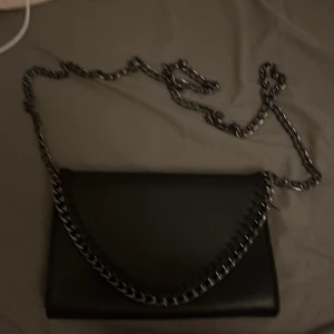 Svart axelväska med kedja - Snygg svart axelväska i slät skinnimitation med kraftig metallkedja som axelrem. Väskan har ett stilrent kuvertformat lock och ett rymligt fack med innerficka. Perfekt för dig som gillar en edgy och clean look.