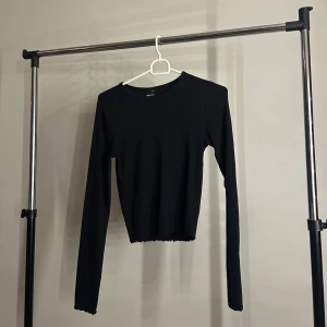 Svart croppad topp Gina Tricot - Svart långärmad croppad topp från Gina Tricot med rund hals och en stor öppen rygg. Toppen är ribbad och har en lätt vågig kant nertill för en extra detalj. Perfekt för dig som vill ha en basic men ändå unik look.